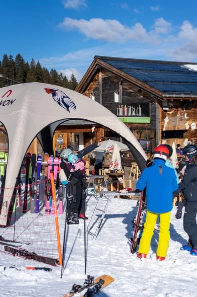 A group of people at a snowy ski resort gathering around an Anavon branded tent, with one person holding a child and various pairs of colorful skis standing in racks nearby.